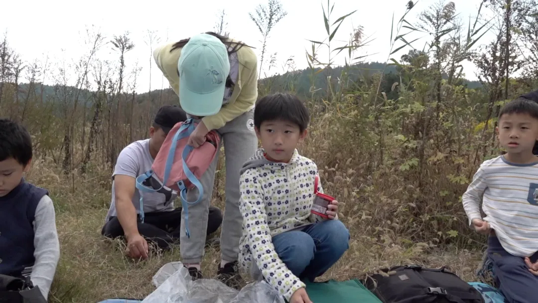 几个孩子和大人在长满高大杂草的荒地上，一名戴帽子的女性整理背包，几个孩子蹲坐或站立在周围。