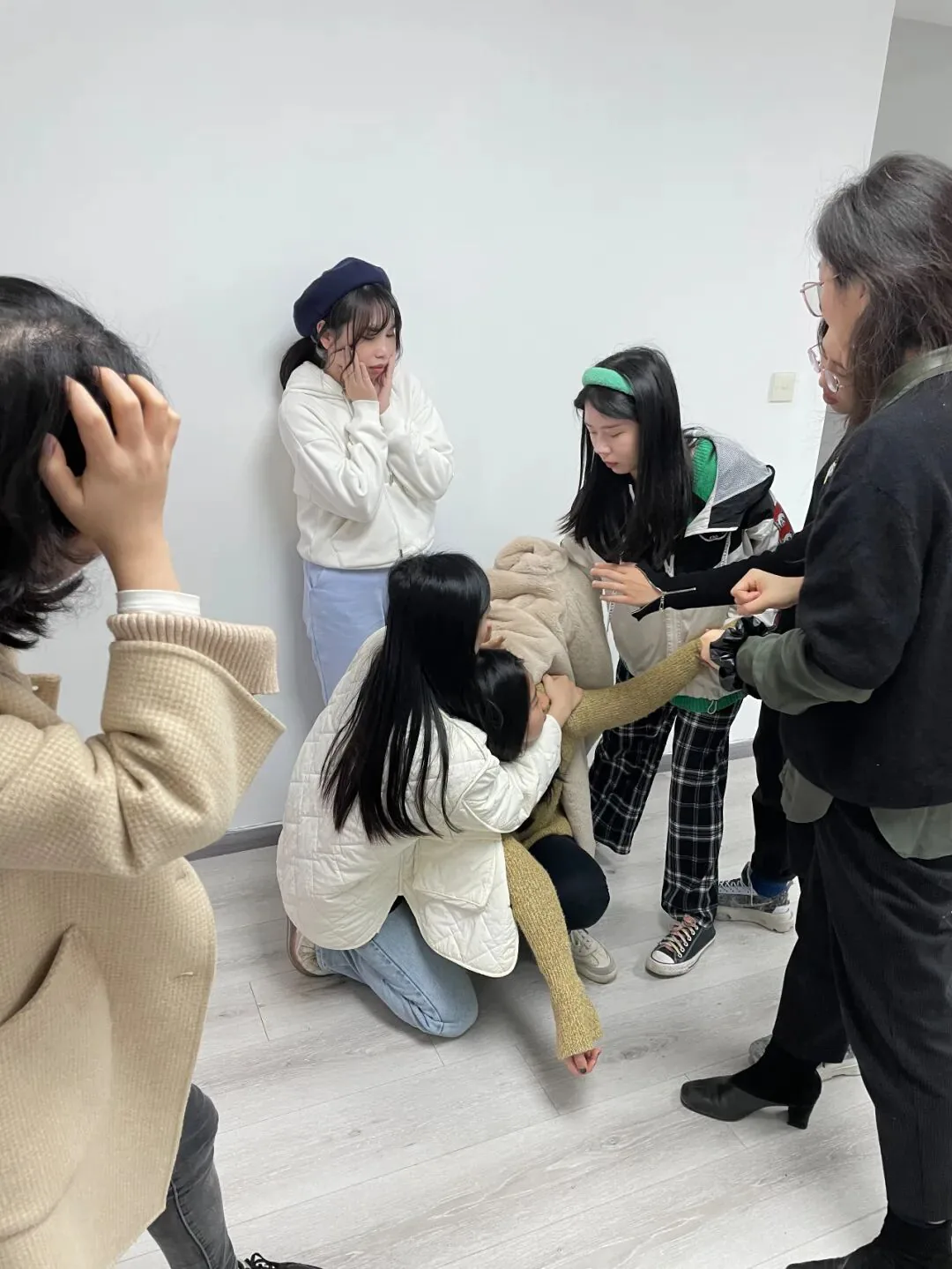 室内几位年轻人正表演情境剧,一人蹲地环抱同伴,其余人做出惊讶、扶额等夸张表情和动作。