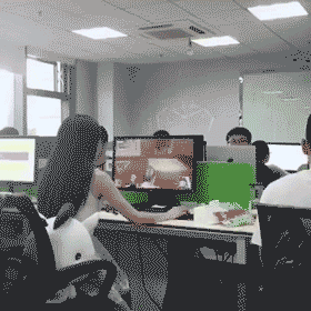 办公室内多名员工在电脑前工作,其中一人的显示器正从视频画面切换为工作文档界面,呈现摸鱼时快速切屏掩饰的场景。