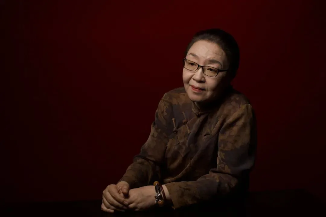 一位戴眼镜、穿深色暗纹上衣的女性坐姿肖像照，双手交叠置于桌面，背景为暗红色。