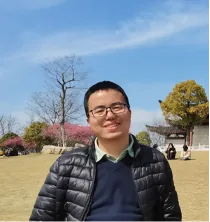 一名戴眼镜、穿黑色羽绒服的男子站在户外草地上留影，背景有樱花树与凉亭。