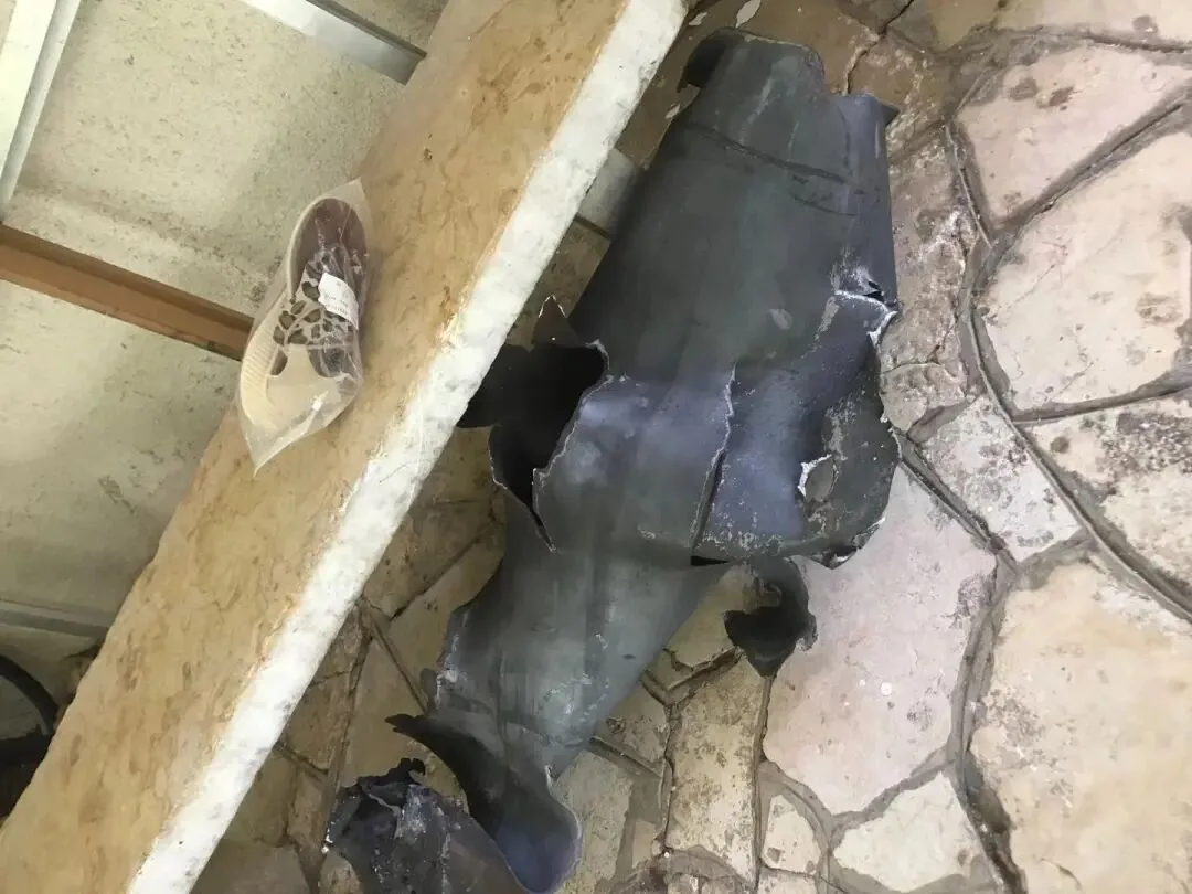 石板地面上有一块撕裂变形的深色金属残片，形似炮弹碎片，一旁台面上放着塑料袋包着的浅色鞋子。