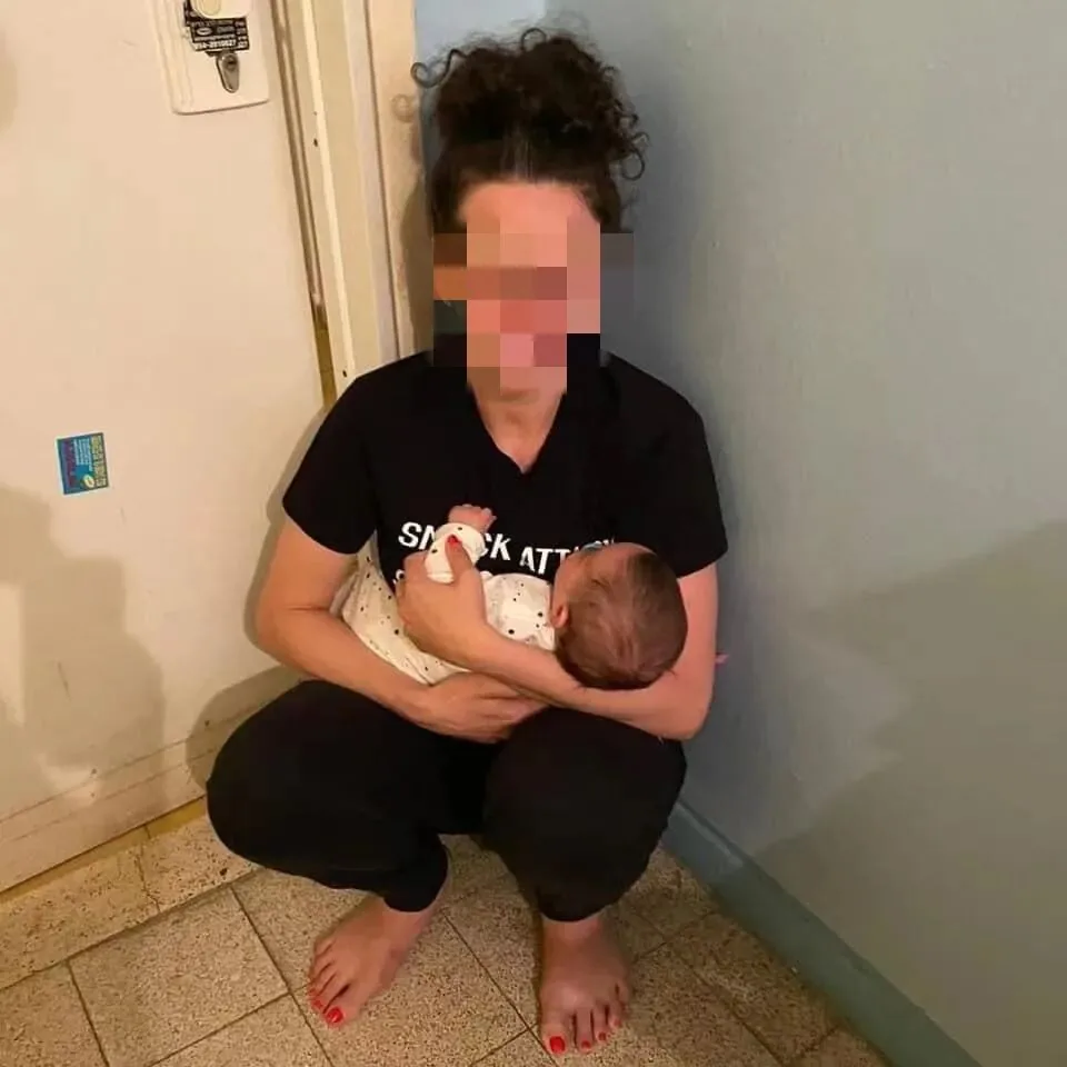 画面中女子面部被打码处理，赤脚蹲在房间墙角地上，双臂抱着一名婴儿，神情紧张地哄着孩子。
