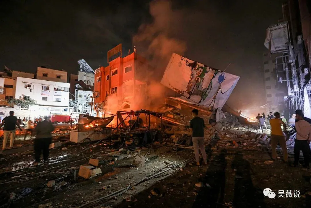 夜间街道上,一栋楼房在空袭后倾斜坍塌并起火冒烟,多人站在废墟旁的黑暗中。