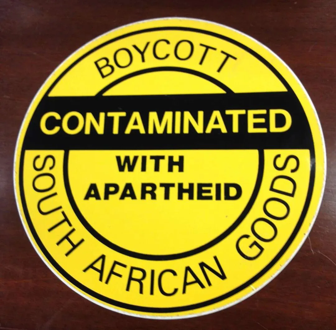 一枚圆形黄黑贴纸置于木质桌面上,环形文字写着 BOYCOTT SOUTH AFRICAN GOODS,中央写着 CONTAMINATED WITH APARTHEID