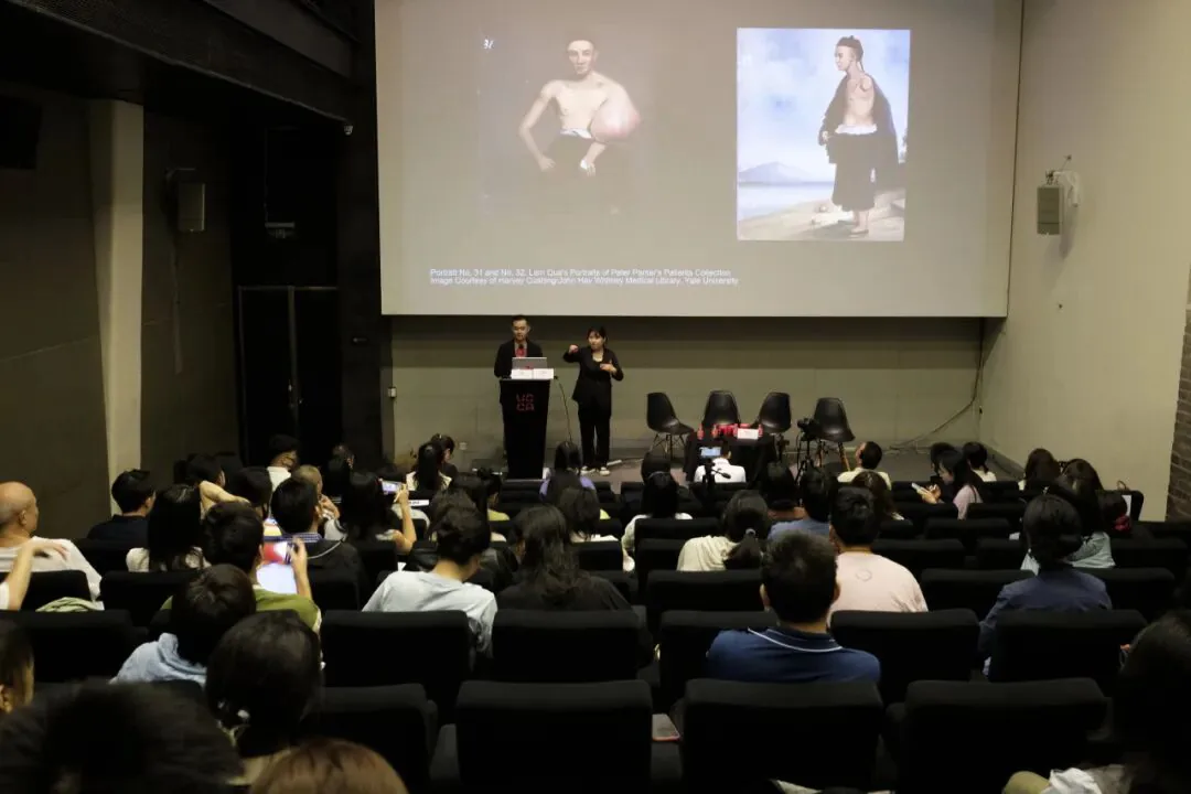 从观众席后方拍摄，大屏幕上展示着此前提到的两幅林官医学肖像画，王超站在讲台前发言，范娇娇在旁打手语，台下坐满观众。