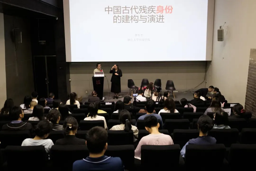 从观众席后方拍摄的报告厅全景，台上李生平站在讲台前发言，手语老师在旁比划，大屏幕显示讲座标题“中国古代残疾身份的建构与演进”，台下坐满观众。