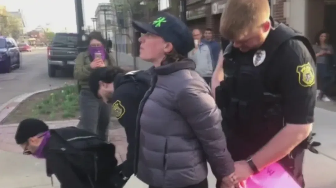 一名戴棒球帽、穿灰色羽绒服的学生被两名校警反手带离,身旁另有一人被按倒在人行道上戴上手铐,周围有围观者举手机拍摄。