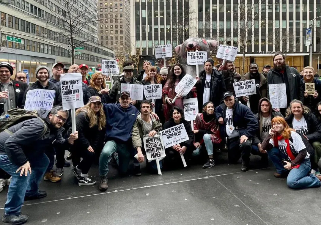 一群人在纽约街头合影,多人举着'New York's Still a Union Town''Alden to News: Drop Dead''New York Needs Its Hometown Paper'等标语牌,其中一人举着一只粉色猪猪偶。