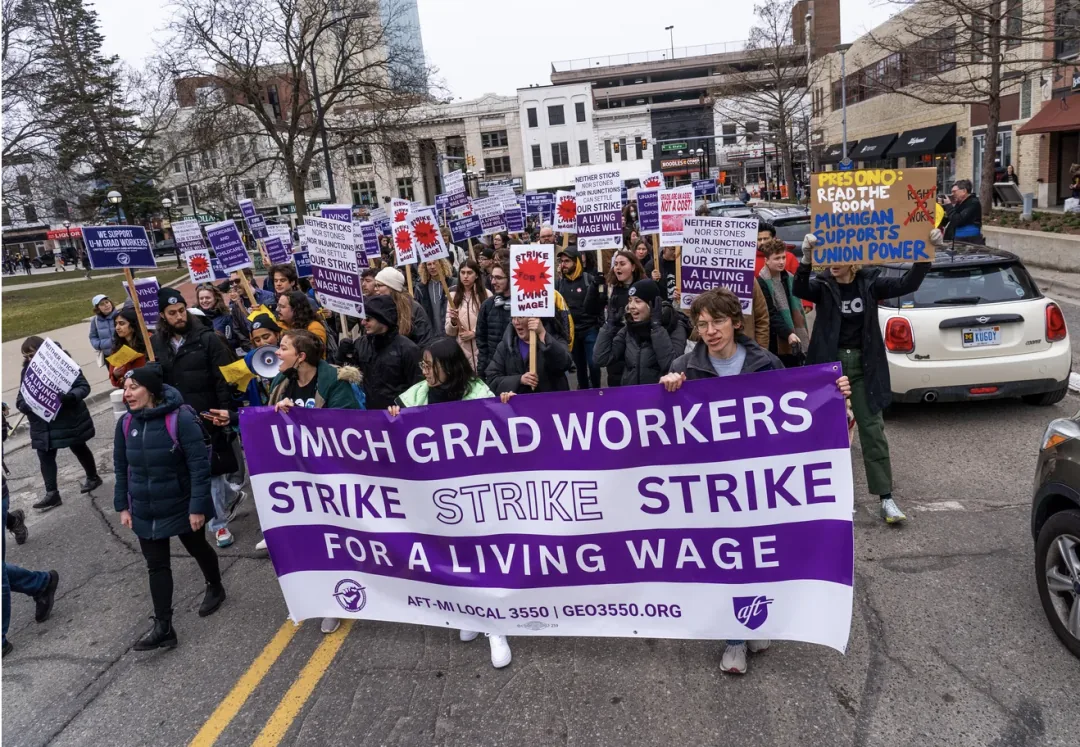 一大群研究生工人在街头游行，前排举着写有'UMICH GRAD WORKERS STRIKE STRIKE STRIKE FOR A LIVING WAGE'的紫白横幅，队伍中许多人举着标语牌，街道两旁是校园建筑。