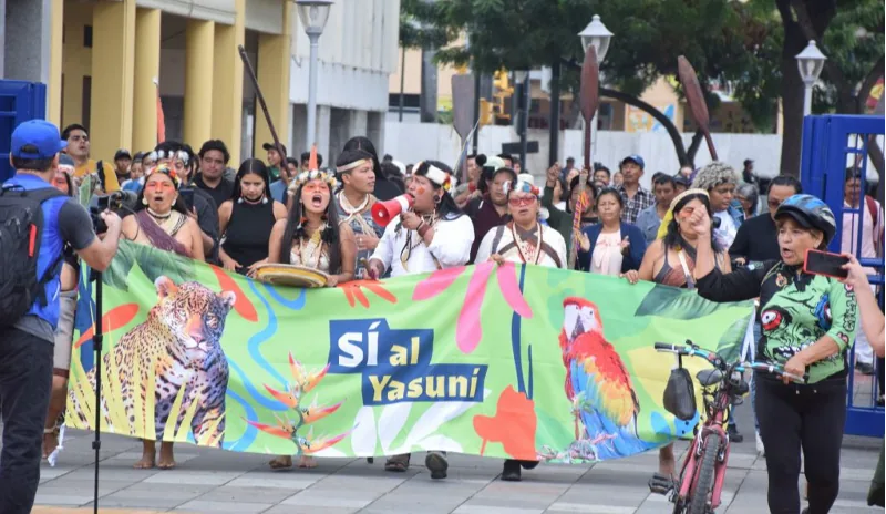 一群头戴传统羽饰的原住民走在街头游行，手持写有“Sí al Yasuní”字样、绘有美洲豹与金刚鹦鹉图案的绿色横幅，两侧民众围观拍照。