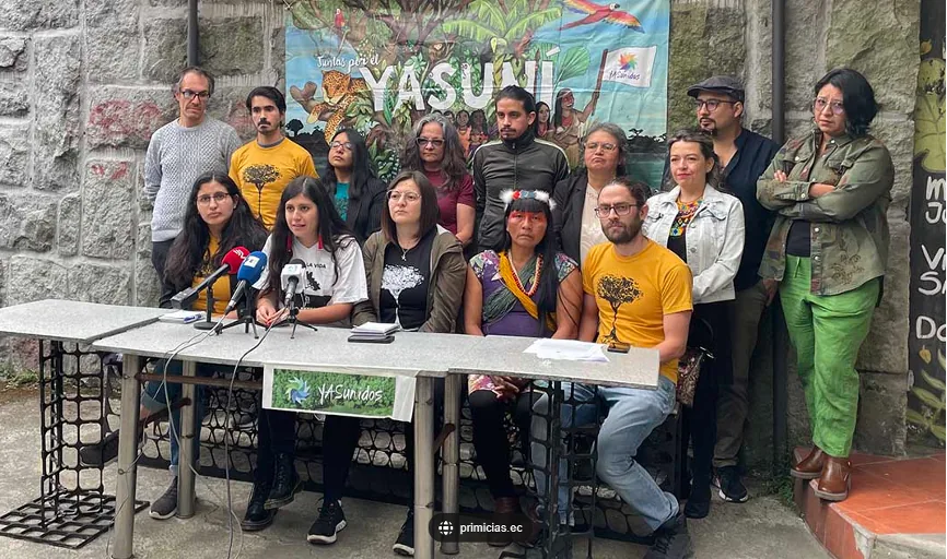 一群身穿黄色“Yasunidos”T恤的运动成员坐在摆有麦克风的桌前召开记者招待会，身后站着支持者，背景是画有雨林与动物的“Yasuní”横幅。