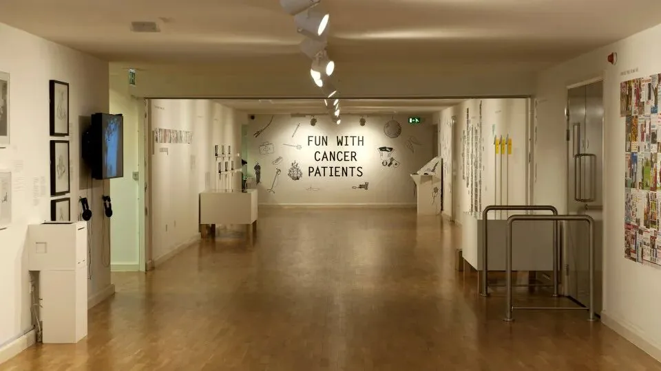 美术馆长廊展览现场，正对面墙上以黑色大字写着“FUN WITH CANCER PATIENTS”，两侧墙面陈列着与治疗相关的手绘图案和展品。