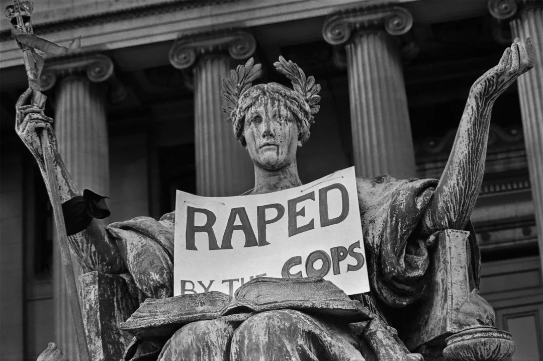 黑白照片,哥伦比亚大学图书馆前的智慧女神雕像怀中被人放置一块牌子,上面写着「RAPED BY THE COPS」。
