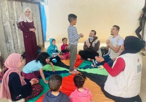 庇护所内几名成人和多名儿童围坐在彩色垫子上做游戏，一名男孩站立在中间，几人戴着口罩或穿着救援机构马甲。