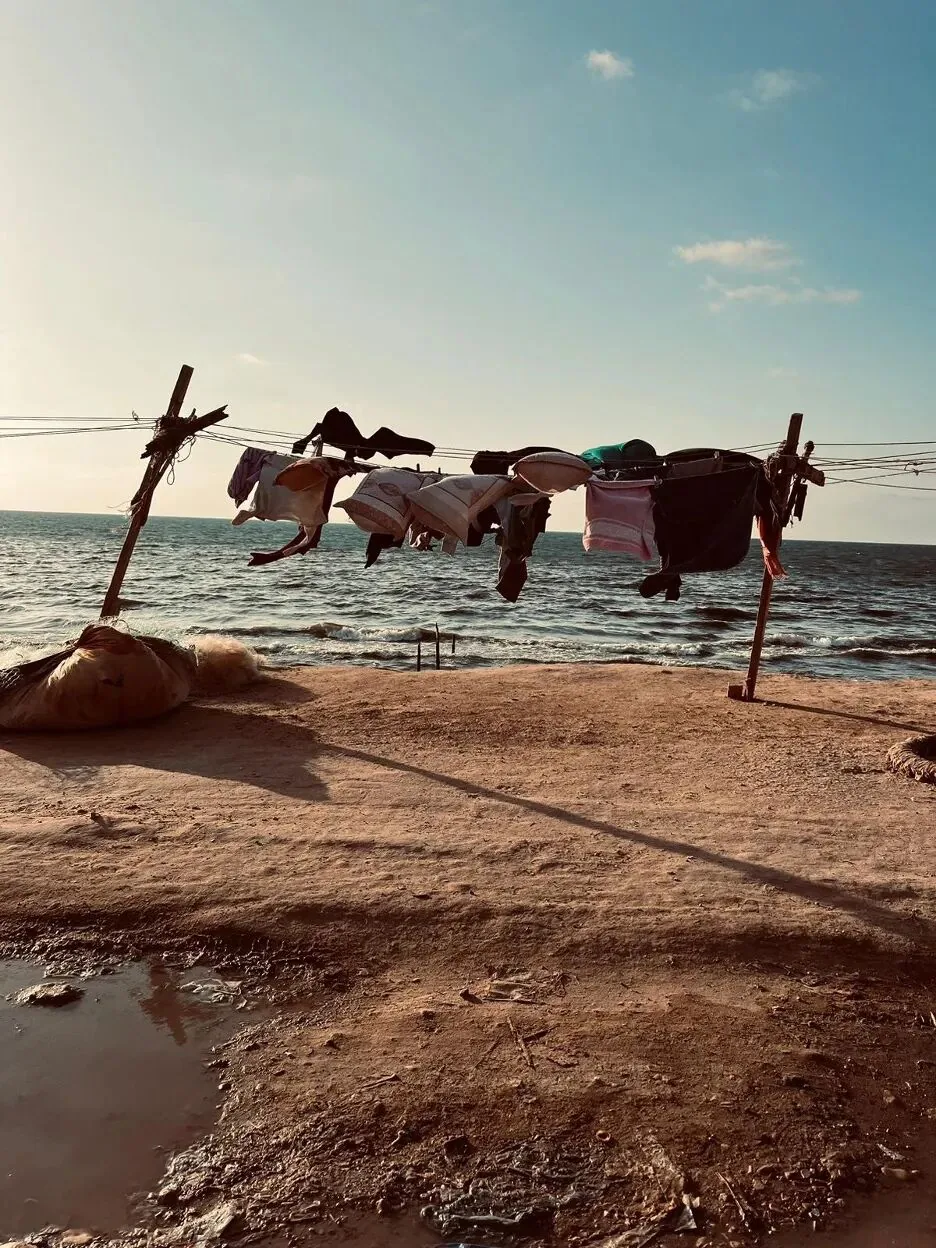 海边沙地上两根木桩间拉起一条绳,晾晒着几件衣物,背景是海面与天空。