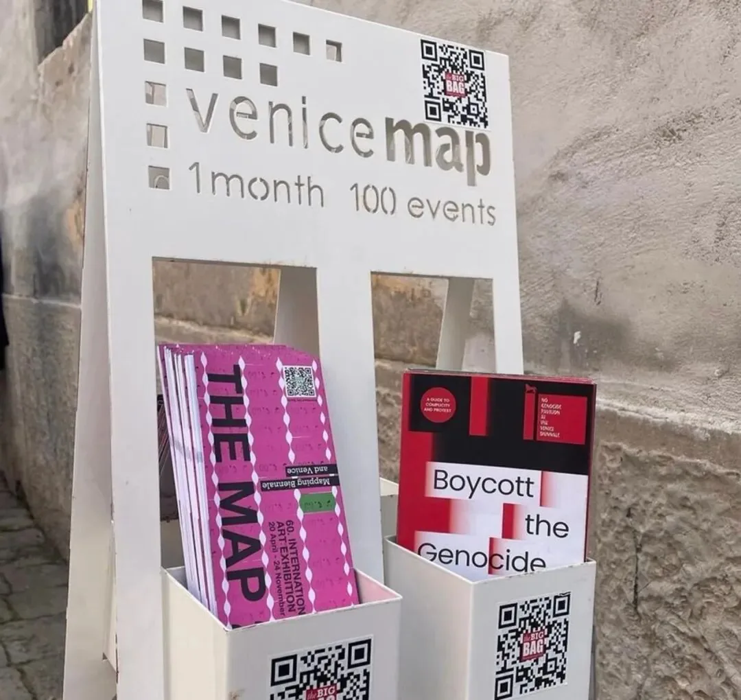 户外宣传架上标有'venice map 1 month 100 events'字样,架内插着粉色'THE MAP'地图册和红黑色封面的'Boycott the Genocide'小册子。