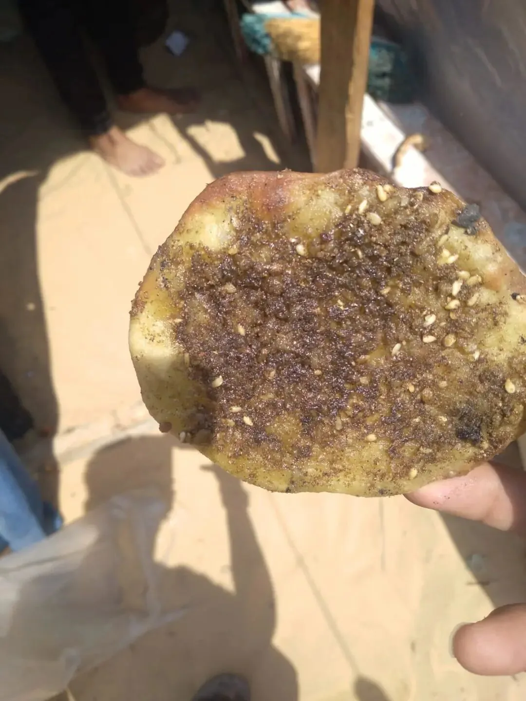 一只手举着一块表面撒满深色调料颗粒的扁面饼，背景为室外地面和一双光脚。