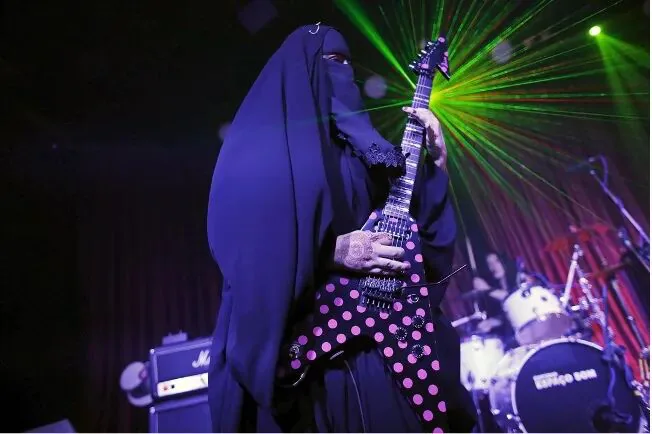 身穿黑色罩袍、蒙面的女性在舞台上弹奏粉色波点电吉他，背景有绿色激光灯与鼓手剪影。