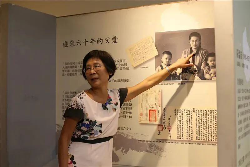 一位女性站在展览板前抬手指向墙上的黑白老照片和文件,展板标题为“迟来六十年的父爱”,照片中是一名男子与两个孩子的合影,旁边陈列着一封手写书信复印件。