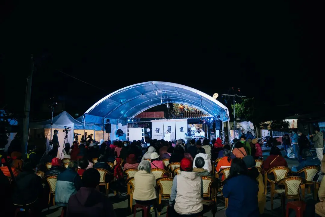 夜晚露天演出现场,观众坐在椅子上围观搭着蓝白色顶棚的舞台,舞台后方是民居和树木。