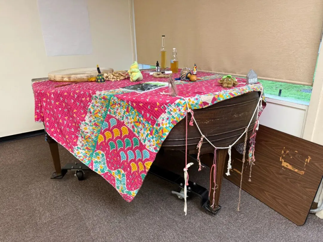 一架旧钢琴上盖着彩色拼布桌布,摆着手鼓、小雕像、瓶装液体等物件,琴身两侧垂挂着多条彩色绳结。