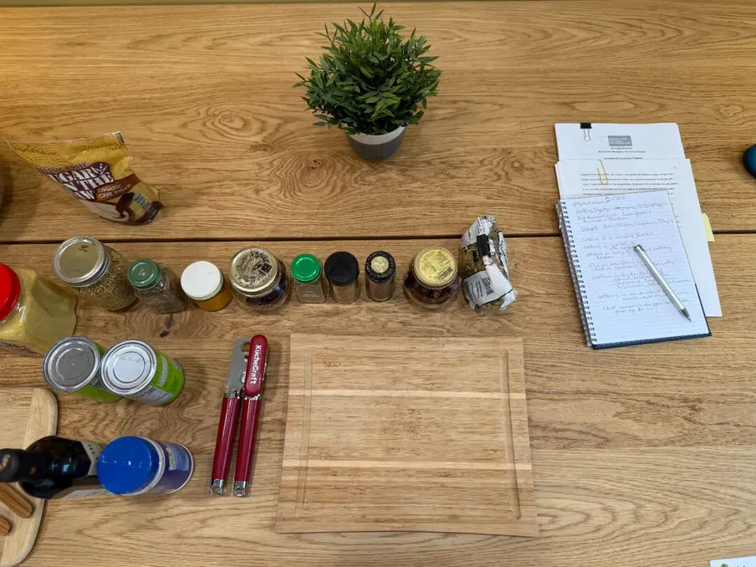 木桌上一排调味料罐、几个罐头、开罐器和一块空案板,桌另一侧放着盆栽绿植和一本摊开的手写笔记本。