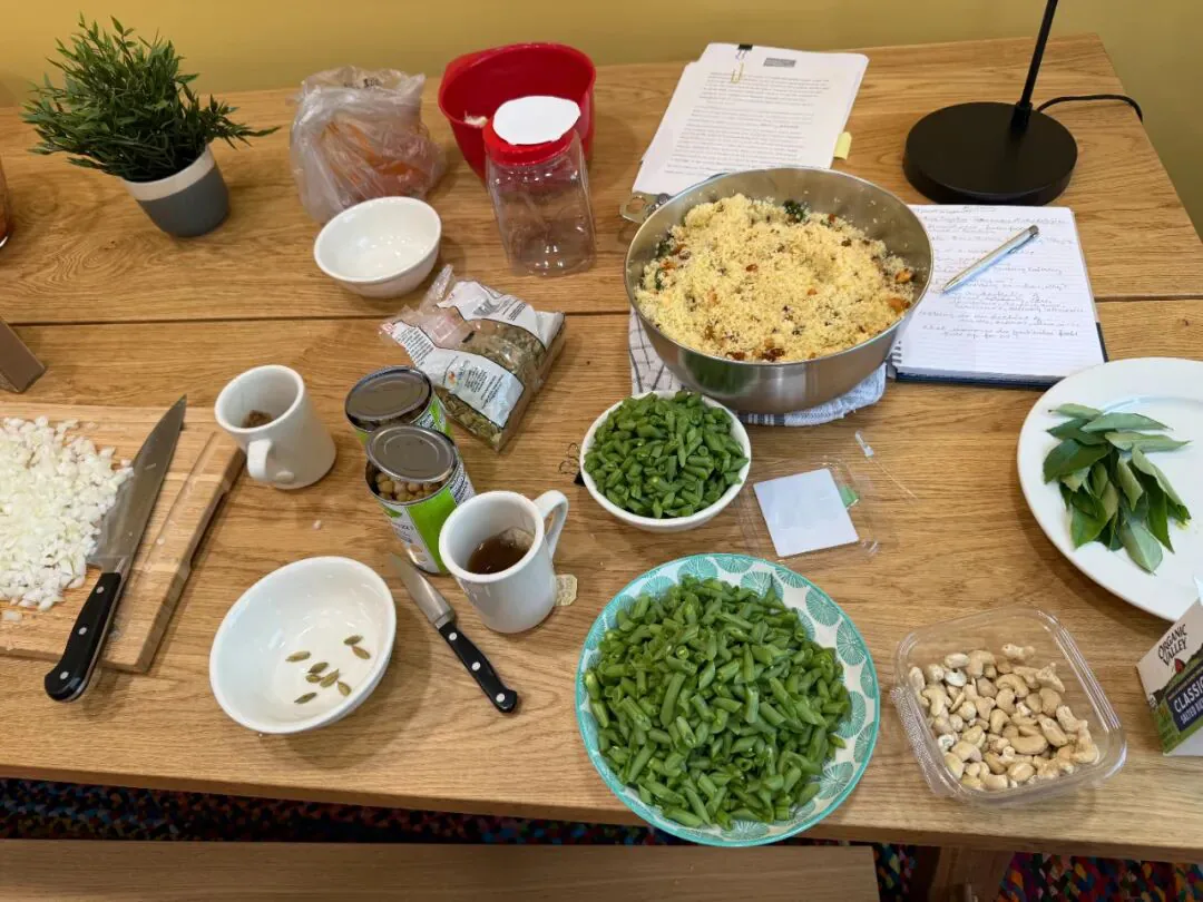 桌上摆着已备好的菜肴:一大锅香料饭、几碗四季豆、腰果和碎奶酪,旁边散放着几只马克杯、刀具和一本笔记本。