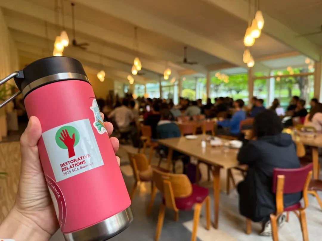 一只手举着粉色保温杯,杯身贴纸印有红色手掌图案和“RESTORATIVE RELATIONS 2025 SCA Biennial”字样,背景是坐满人用餐的集体餐厅。