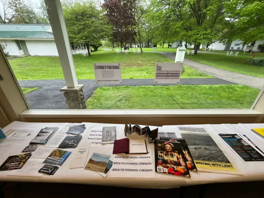 窗边长桌上摆满各种小册子和杂志,多本标注“Bureau for Experimental Ethnography”,其中一本封面写着“Writing with Light”;窗外草坪上立着写有“EXPANDED PUBLISHING”的展牌。