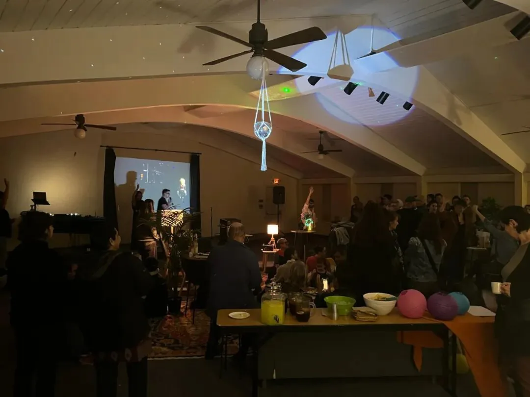 室内夜间聚会场景,彩色射灯投影在天花板上,人群在放映屏幕前跳舞聚集,前景桌上摆着饮料罐和几只彩色气球。