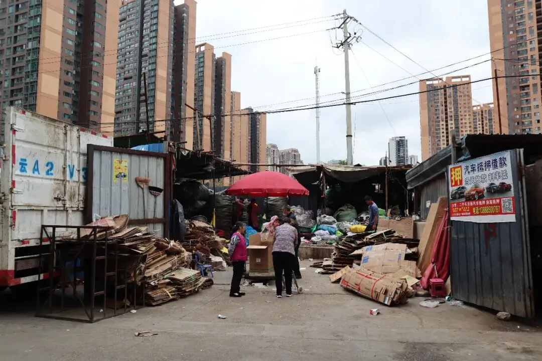 回收站堆满纸板和杂物,几人在红色遮阳伞下交谈,四周是简易棚屋和货柜车,背后高楼林立。