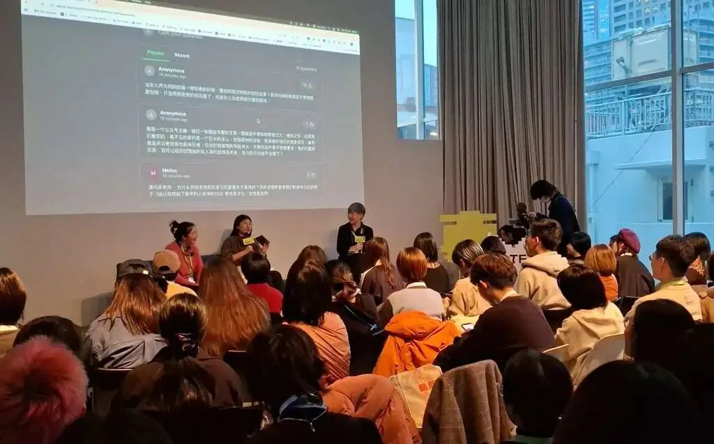 三位讲者站在台前对谈，身后大屏幕展示听众匿名提交的中文提问和留言，台下观众坐满整个空间，落地窗外可见城市高楼。