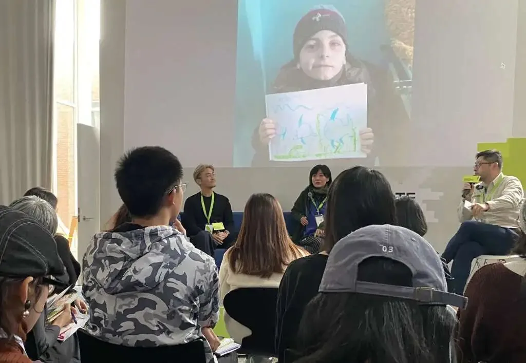 大屏幕播放一名戴毛线帽的孩子举着自己手绘图画的视频画面，台前几位讲者围坐对谈，台下观众围坐聆听。