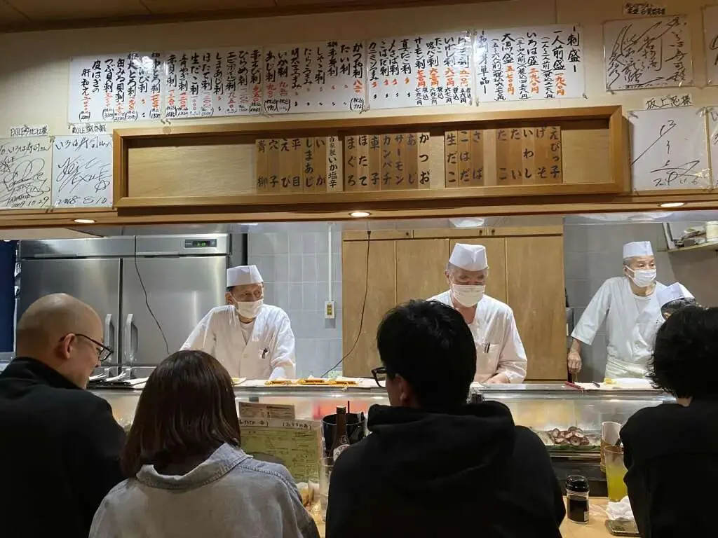 一家寿司店柜台内三位戴口罩的男性厨师在准备食物，头顶挂满手写日文菜单木牌，柜台前坐着几位顾客。