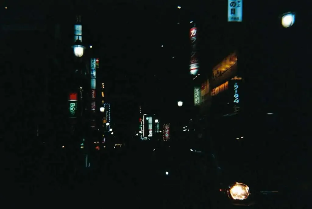 夜晚的东京街道，两侧竖立着多块发光的日文招牌，昏暗中一辆汽车的车头灯在画面右下角亮起。