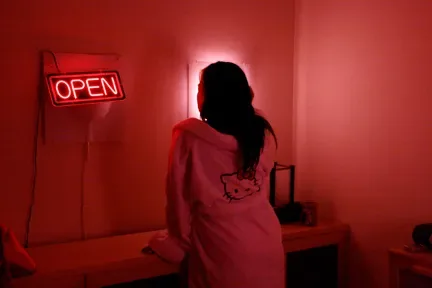 红色灯光笼罩的房间里，一人背对镜头站立，身穿印有凯蒂猫轮廓图案的连帽衫，墙上挂着亮着的“OPEN”霓虹灯牌