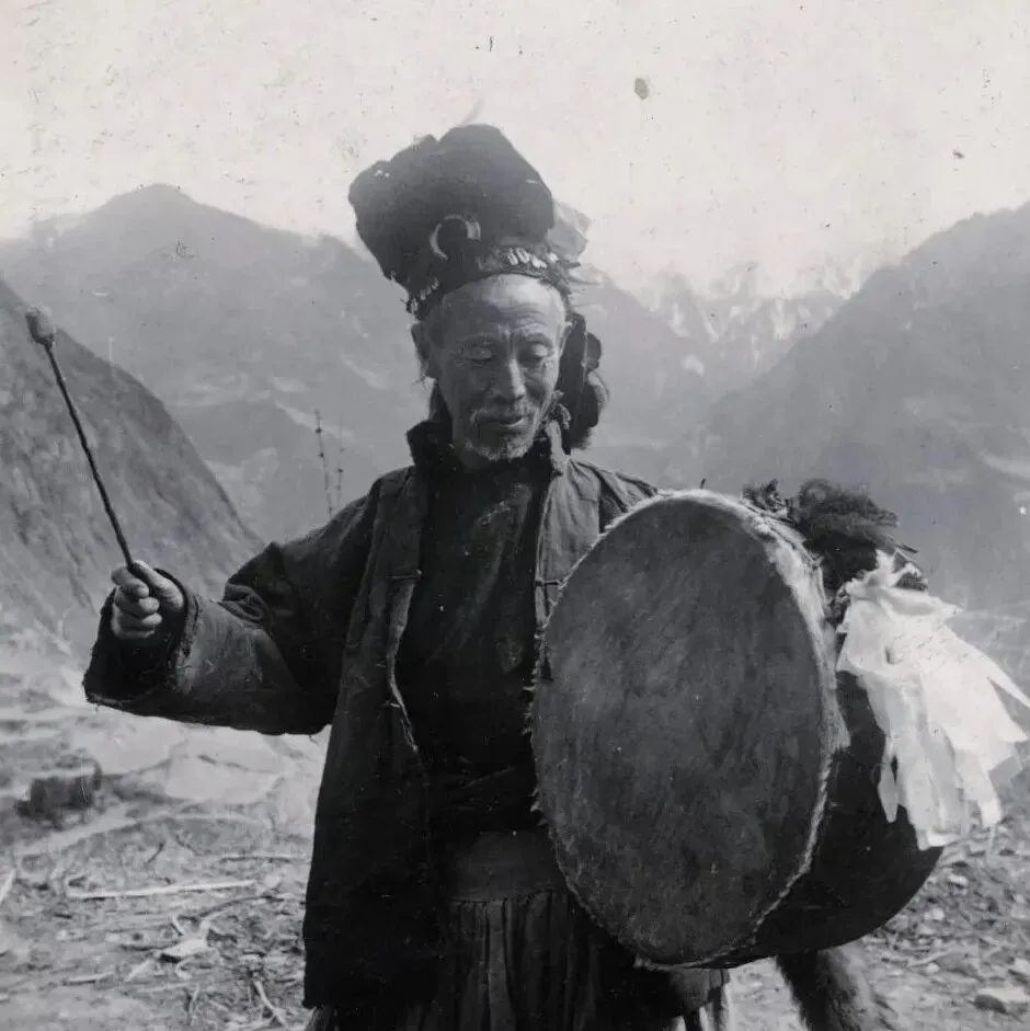 一位头戴羽饰头饰的老年男子（端公）站在群山间空地上，一手持鼓槌一手持大鼓，正在敲鼓作法。