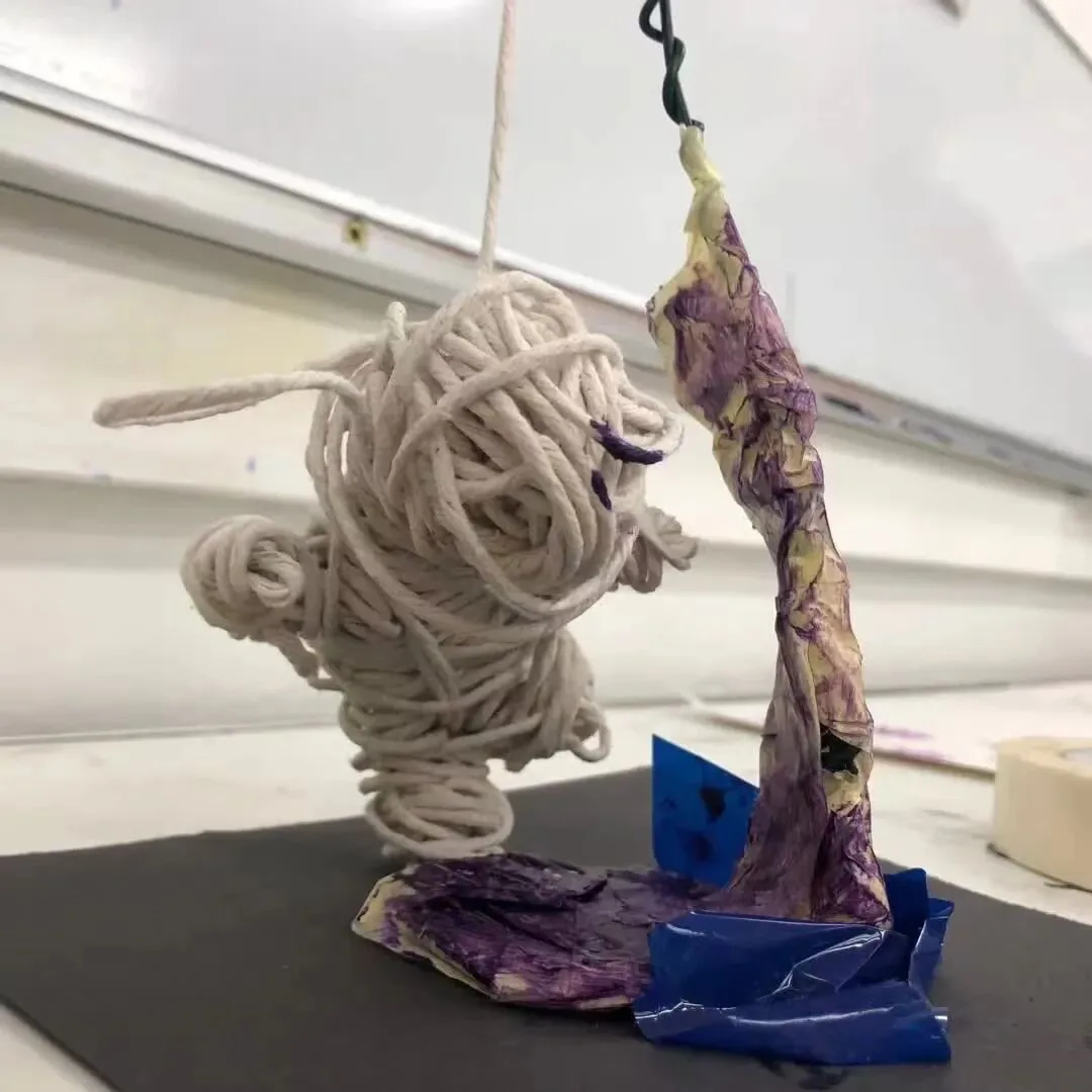 用白色棉绳缠绕成的人形雕塑立于深色台面,一旁立着染成紫色的纸质柱状造型,底座为蓝色黏土。