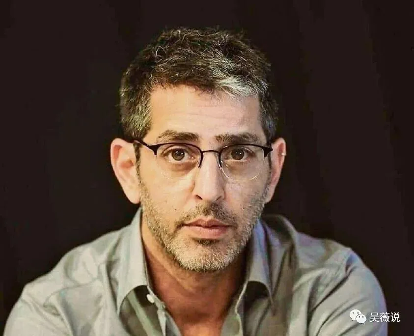 罗伊·伊丹戴眼镜、灰白短发,身穿灰色衬衫,暗色背景下的正面肖像照。