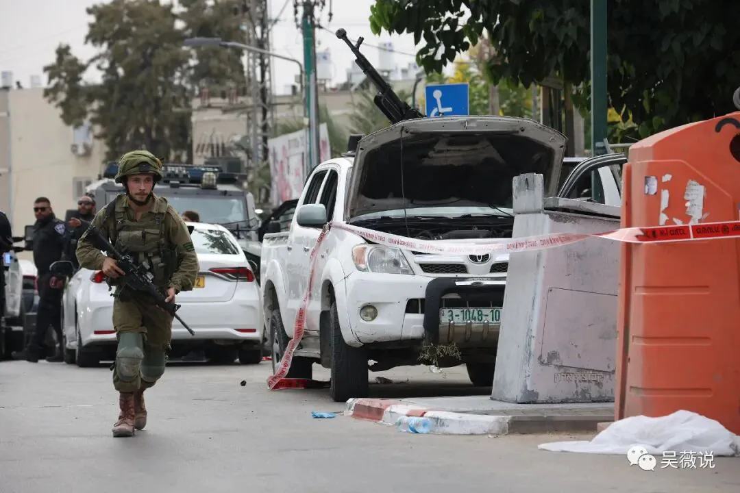 街道上,一名持枪的以色列士兵跑步经过一辆车顶架有机枪、引擎盖打开的皮卡车,车辆已被警戒带围起。