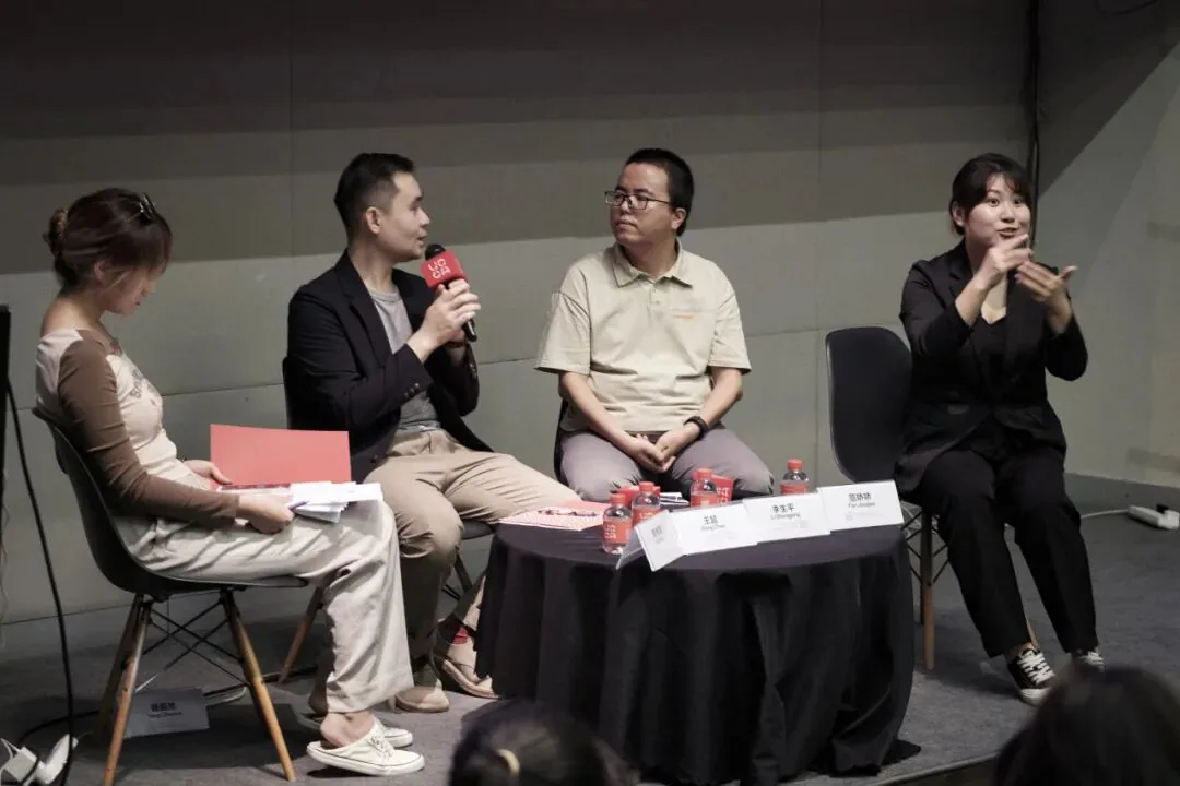 四人并排坐在台上的小圆桌旁,王超手持话筒发言,范娇娇在最右侧打手语,桌上摆放着姓名牌和瓶装水,呈现对谈现场氛围。