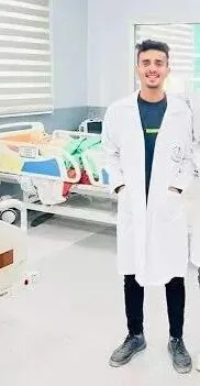 身穿白大褂的年轻男子在病房里微笑站立,身后是病床和医疗设备。