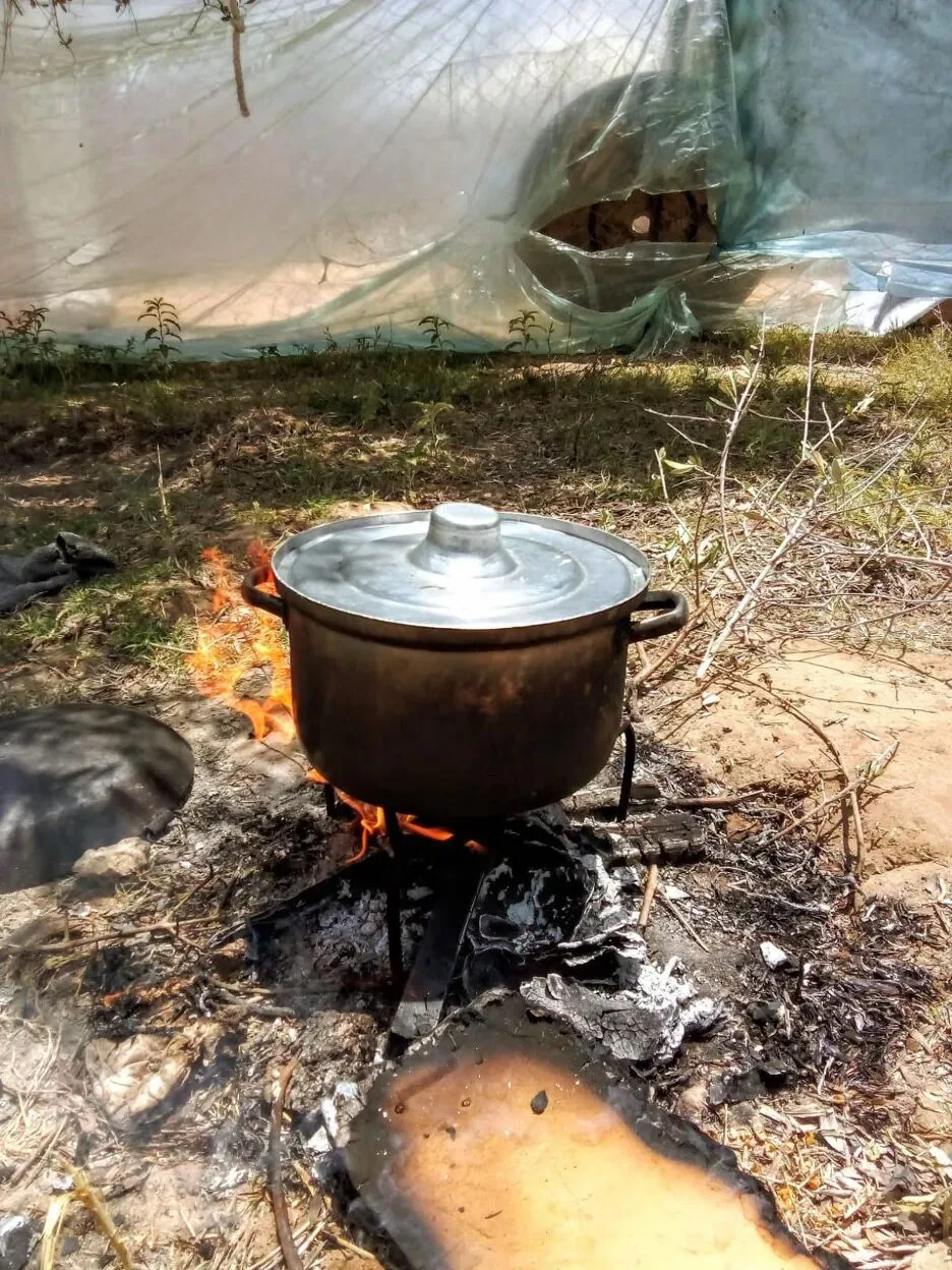 露天空地上,一口黑色铁锅架在柴火上炖煮,背景是塑料布搭起的简易帐篷。