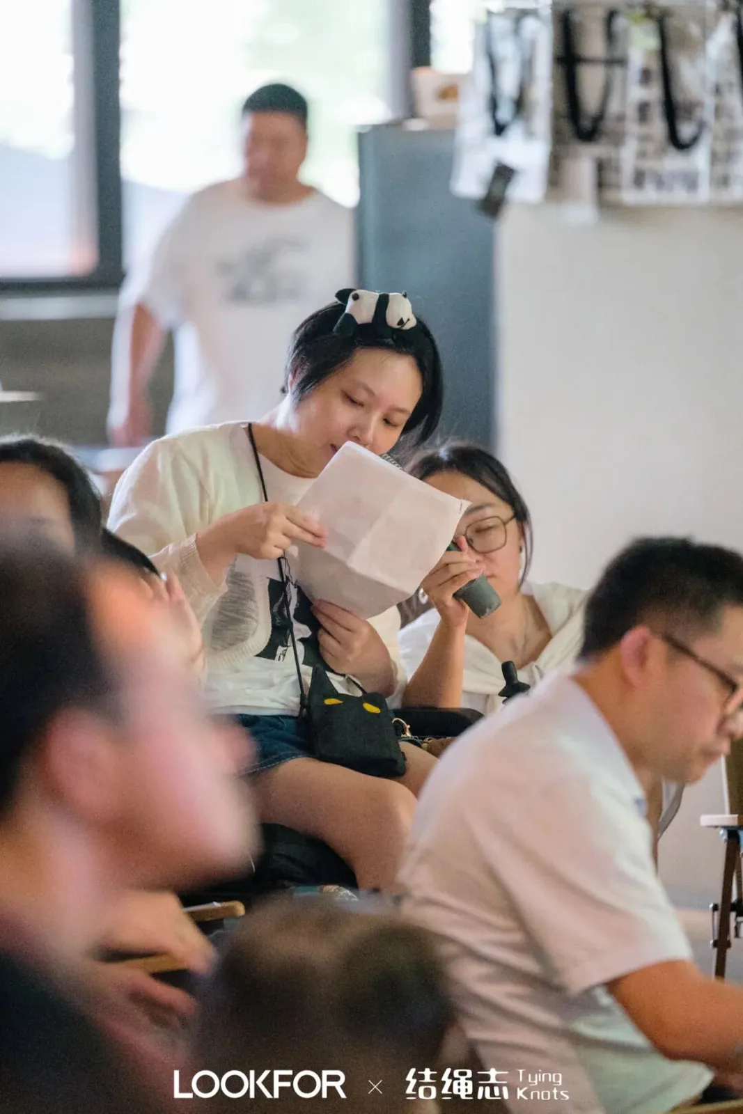 沈妍头戴熊猫耳发箍,坐着举起一张纸凑近眼前阅读,身旁一女子捂嘴倾听,背景有人经过。