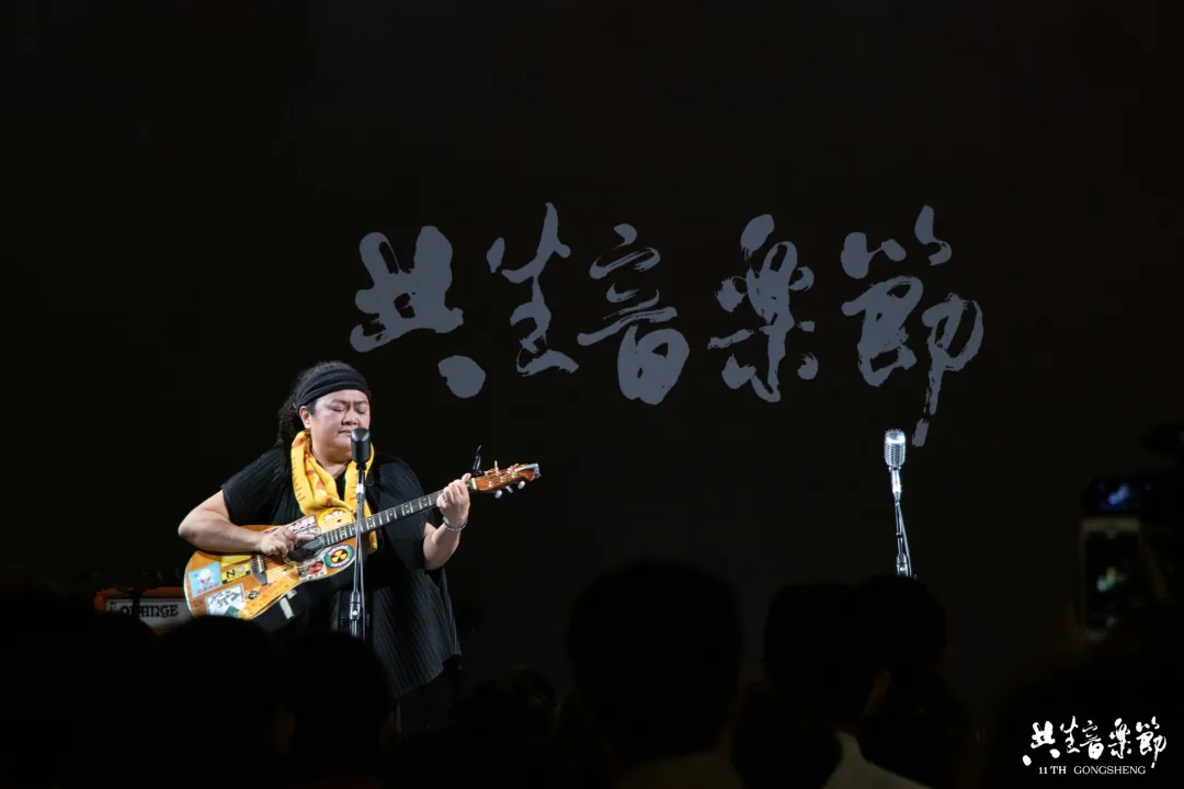 一位戴头巾、系彩色围巾的女性在台上抱着吉他弹唱，身后暗色背景写着“共生音乐节”字样。