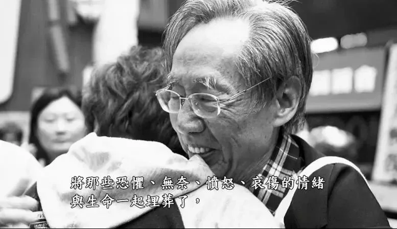 纪录片截图，黑白画面中戴眼镜的老年男性微笑着拥抱他人，画面字幕为“将那些恐惧、无奈、愤怒、哀伤的情绪与生命一起埋葬了，”