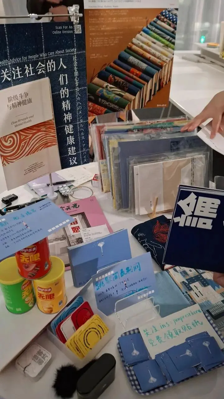 市集摊位桌面摆满自制小志、明信片和铁盒零食，海报副标题写着“阶级斗争与精神健康”（Class Struggle and Mental Health），旁桌还有蓝色手写卡片。
