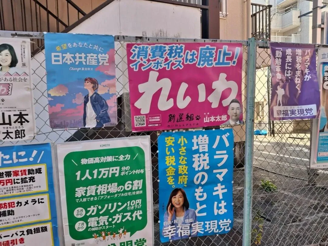 一面铁丝网围栏上贴满日本政党竞选海报，可见“日本共产党”、“れいわ”（配山本太郎头像，标语“消费税・発票废止！”）、“幸福実現党”等，背景是居民楼。