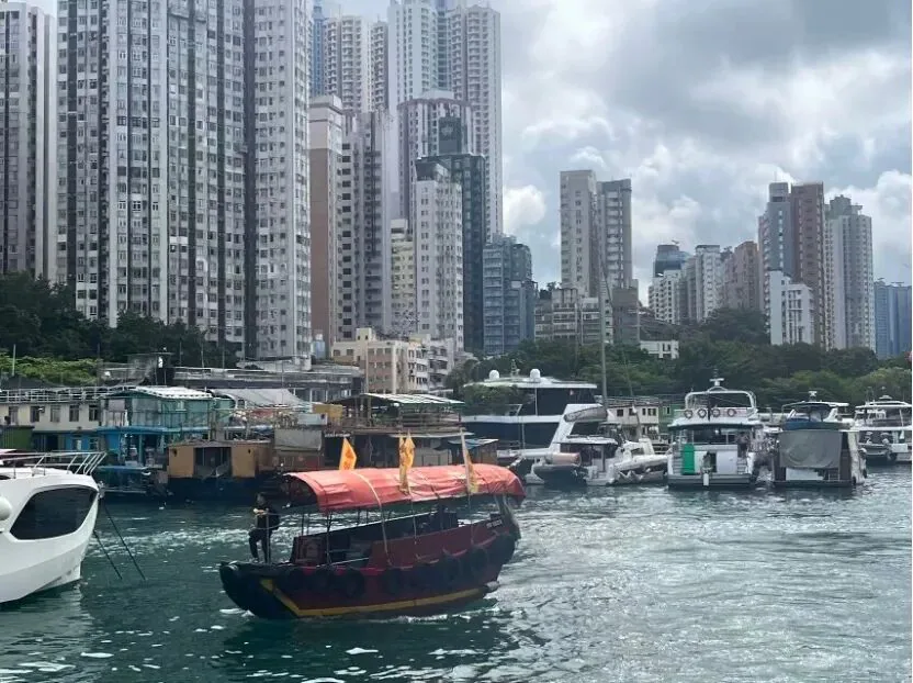 一艘红顶传统渔船驶过避风塘水面,背景是密集的高层住宅楼,岸边停靠着多艘白色游艇与旧式渔排屋